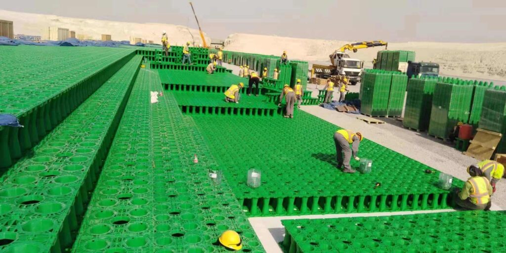 Workers installing large-scale green geocellular rainwater harvesting modules at a Middle East construction site with desert terrain and excavator in background
