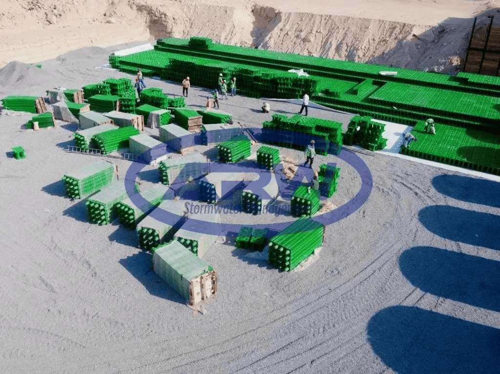 Módulos geocelulares verdes para aguas pluviales apilados en una zona de preparación de obra en Kuwait; trabajadores preparan su instalación sobre una subbase de grava compactada