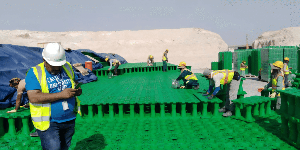 Site engineer supervising geocellular stormwater module installation at Dubai logistics park with workers assembling green polypropylene crates on desert sand base
